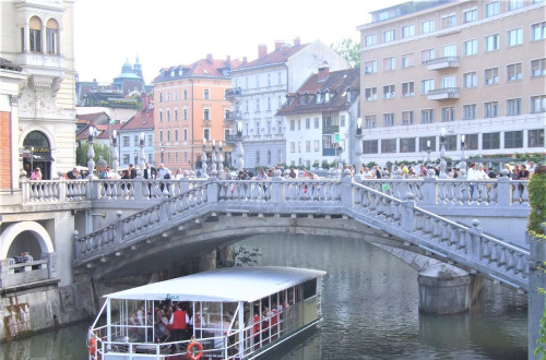 Na ladjici na reki Ljubljanici igra pihalna godba, ljudje pa jo z mostu opazujejo in poslusajo.