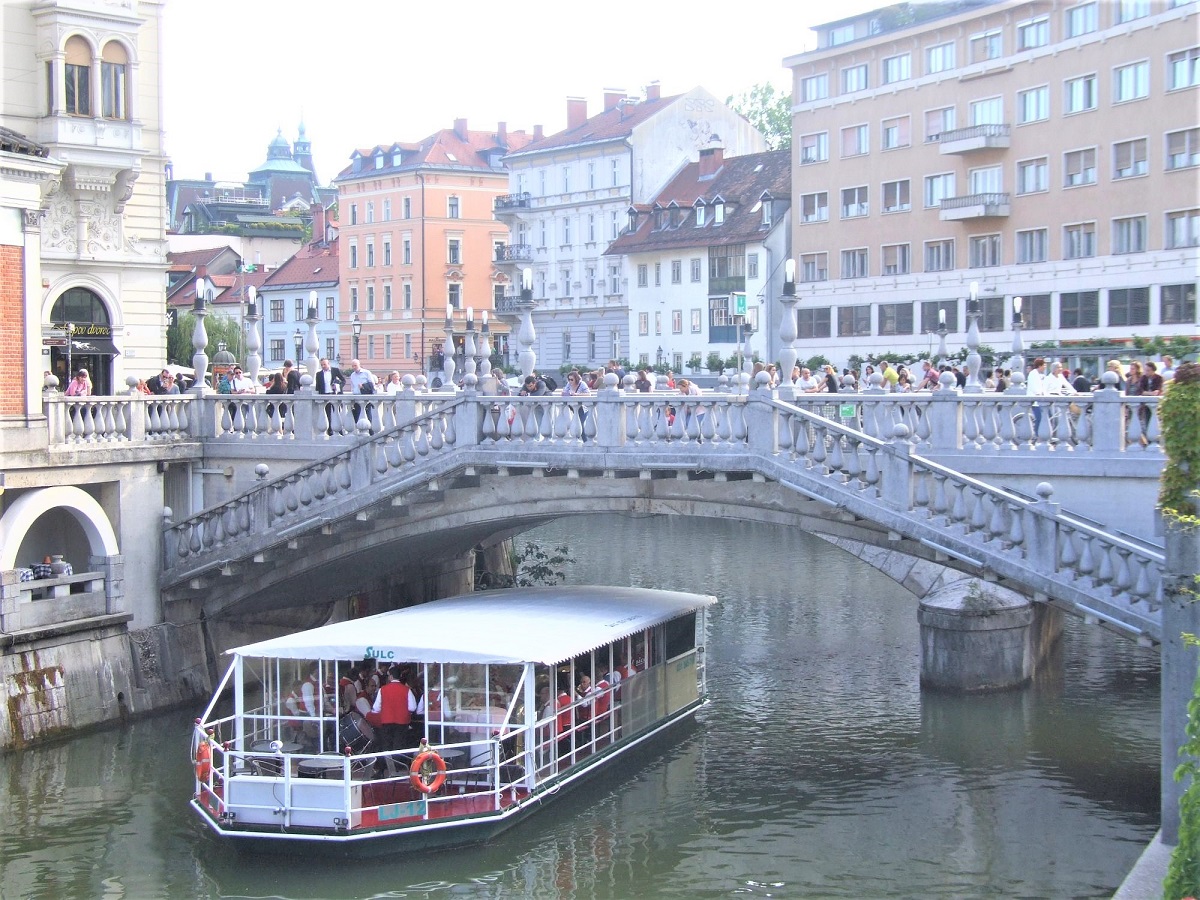 Na ladjici na reki Ljubljanici igra pihalna godba, ljudje pa jo z mostu opazujejo in poslusajo.