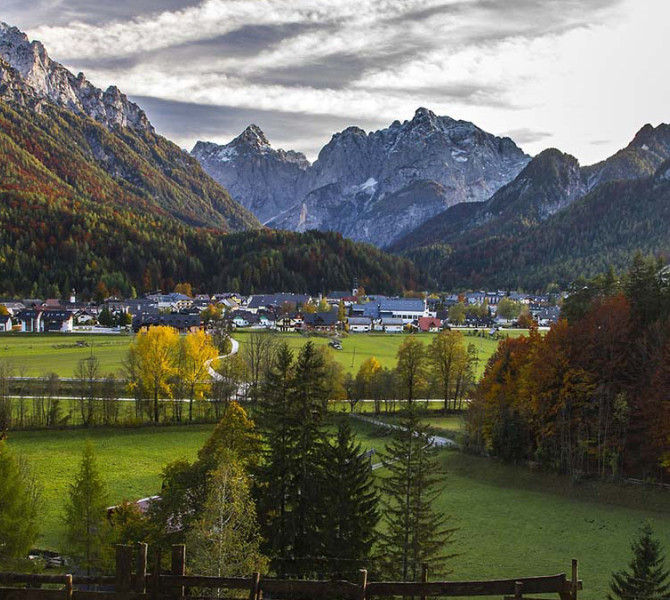 Une vue panoramique d'un village de montagne dans une vallée, entouré de forêts aux couleurs automnales et de puissants sommets enneigés en arrière-plan.