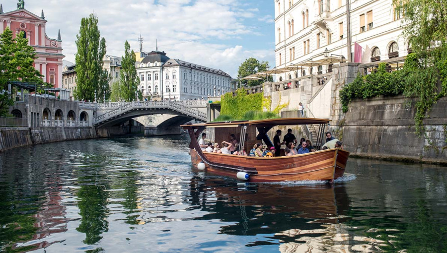 Ljudje plujejo na leseni barki po reki Ljubljanici, v ozadju stavbe v srediscu Ljubljane.