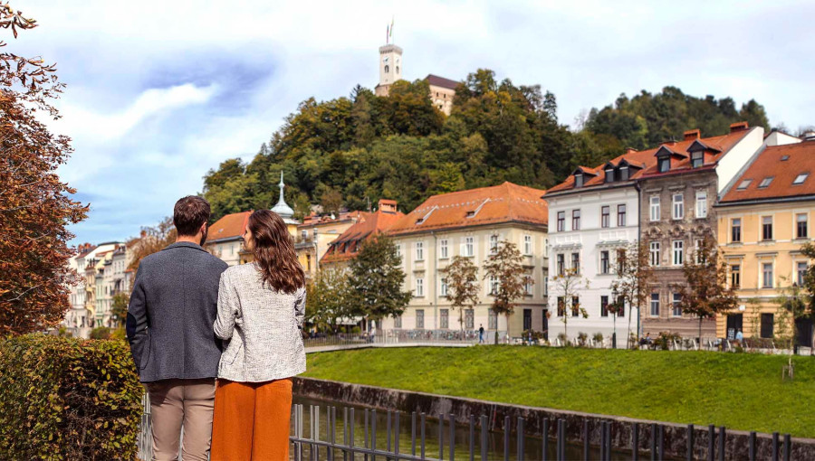 Zaljubljen par stoji na nabrežju Ljubljanice, v ozadju Ljubljanski grad.