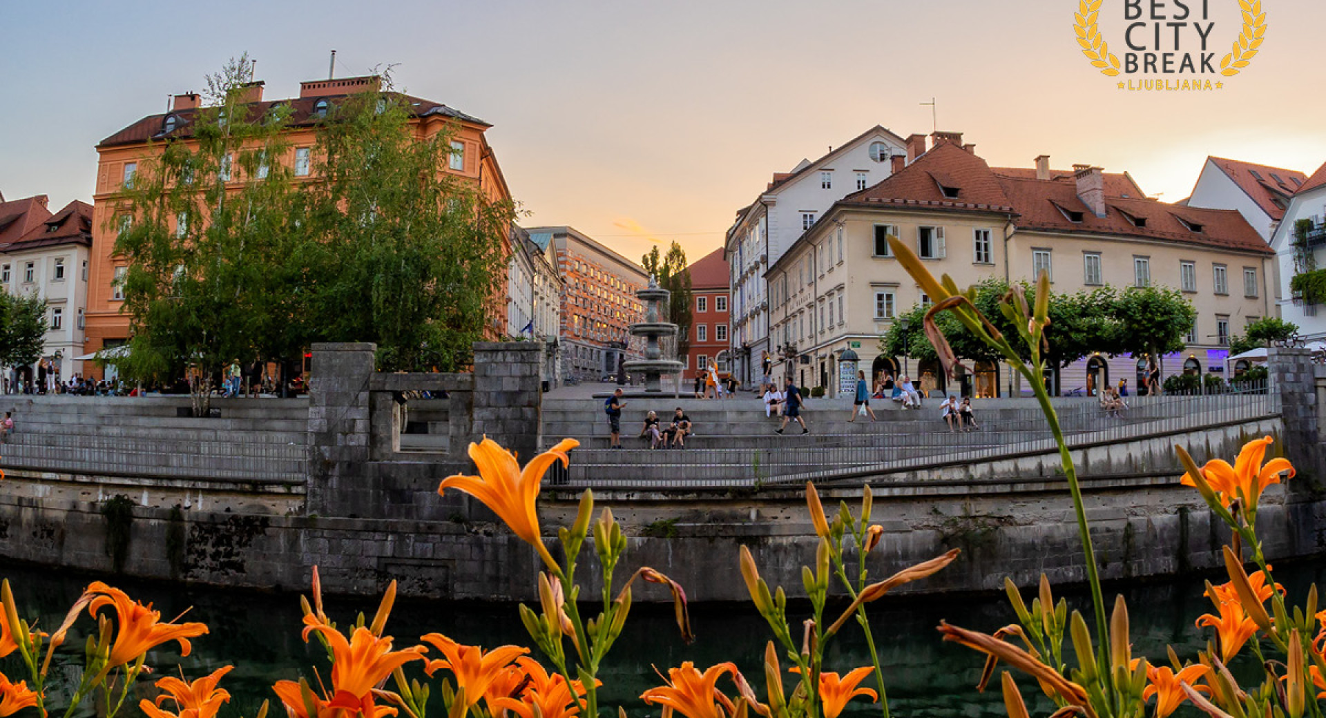 A charming city centre with a fountain. Orange lilies on the river front. People sitting on the river bank.