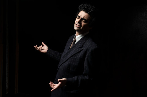 Moski v crnem suknjicu izvaja opero, za njim pa je crno ozadje.