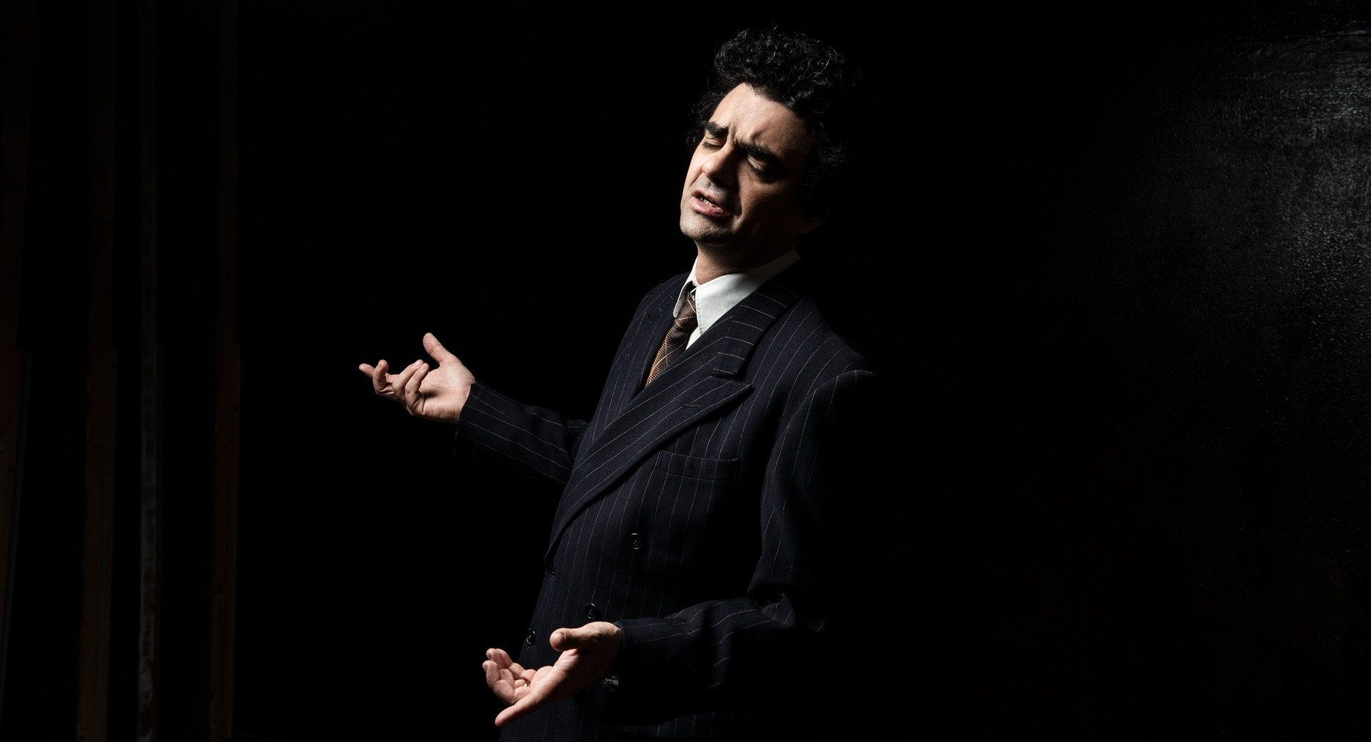 Moski v crnem suknjicu izvaja opero, za njim pa je crno ozadje.