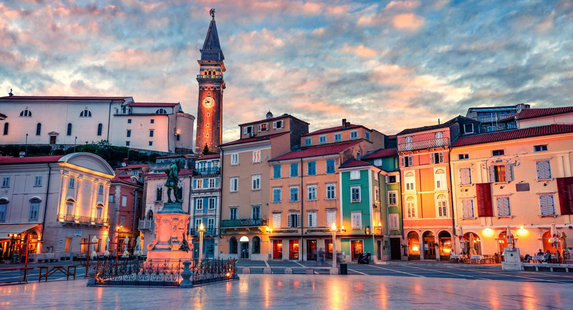 Ein steinerner Platz mit beleuchteten bunten Fassaden, einer Statue und einem Glockenturm in der Abenddämmerung.