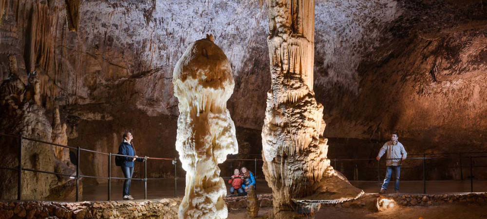 A large dripstone cave with massive stalagmites and stalactites and visitors walking along a secured path with a railing.