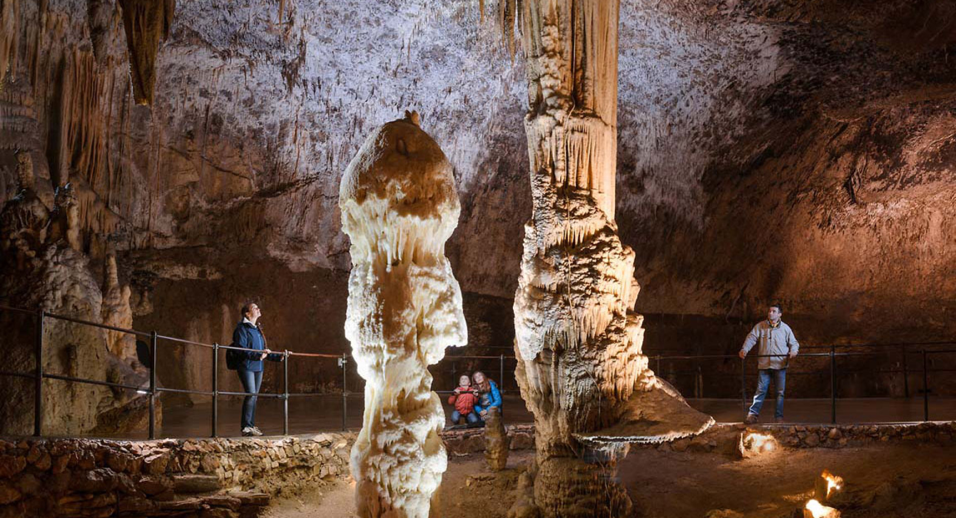 Notranjost Postojnske jame z obiskovalci na mostu. V ospredju osvetljena stalagmita.