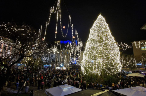 Pogled na osrednji mestni trg z daljave zvecer. V srediscu smreka z rumenimi lucmi, okoli nje z rumenimi lucmi okrasene ulice in drevesa.