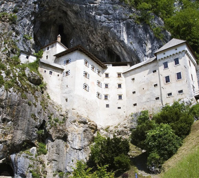 Predjamski grad je vpet v skalno steno nad zeleno pokrajino pod mogocno jamsko odprtino.
