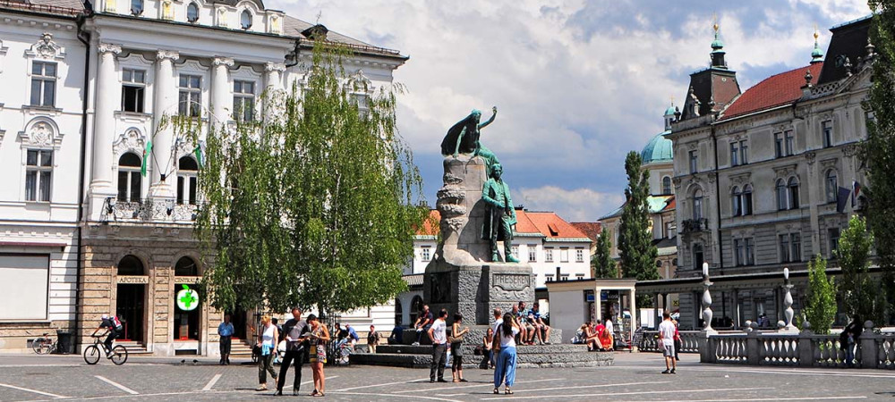 Na zivahnem trgu se nahaja velik kip figure z dvignjeno roko, ki stoji na kamnitem podstavku. V ozadju so stavbe in drevesa, ljudje pa se pod delno oblacnim nebom sprehajajo in zbirajo v blizini kipa.
