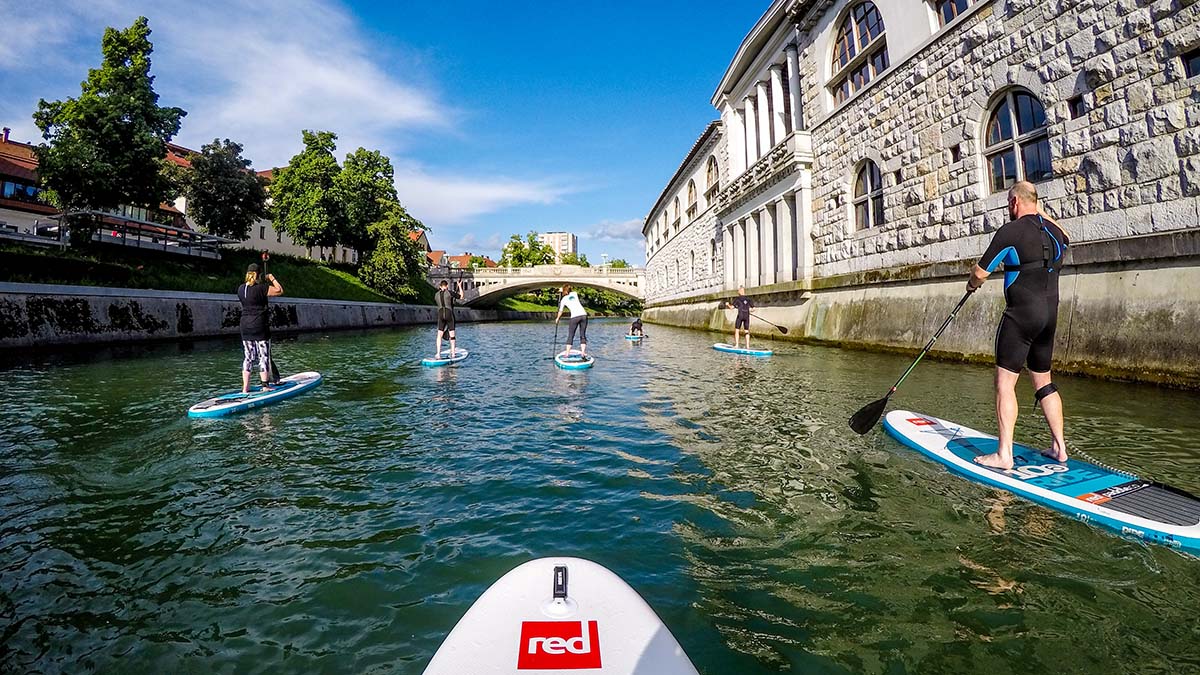 Five paddleboarders on the Ljubljanica River. On the right side is the Plečnik Market. On the left side is the riverbank.