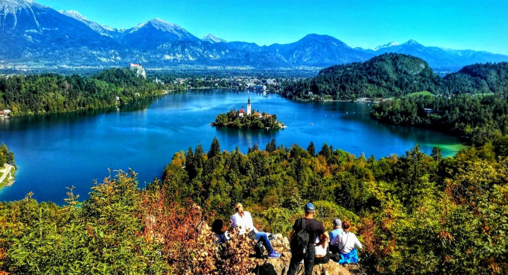 Aussicht von einem Hügel auf den Bleder See mit der Insel, vier Wanderer stehen auf dem Hügel, Berge im Hintergrund.
