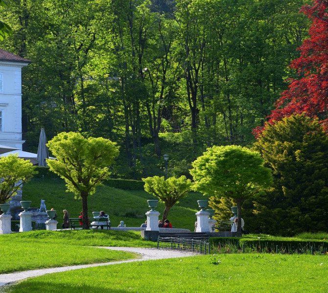 Spomladanski park Tivoli z zivahno zelenico, drevoredom in Gradom Tivoli v ozadju, umescenim v gozdnato pobocje.