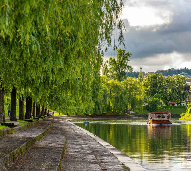 Lesena rjava barka pluje po reki ob zelenih nabrezjih s sprehajaliscem.