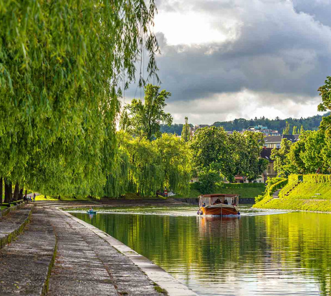 Lesena rjava barka pluje po reki ob zelenih nabrezjih s sprehajaliscem.