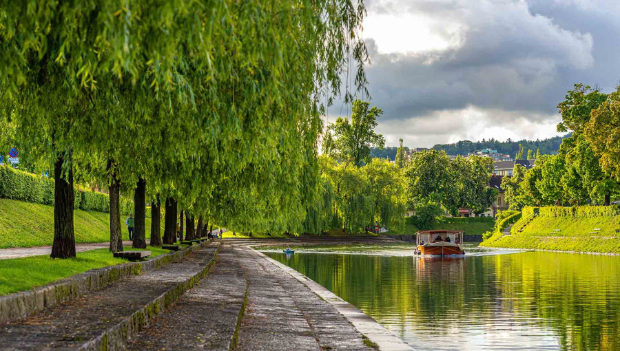 A brown wooden boat is sailing along the river beside green riverbanks with a promenade.