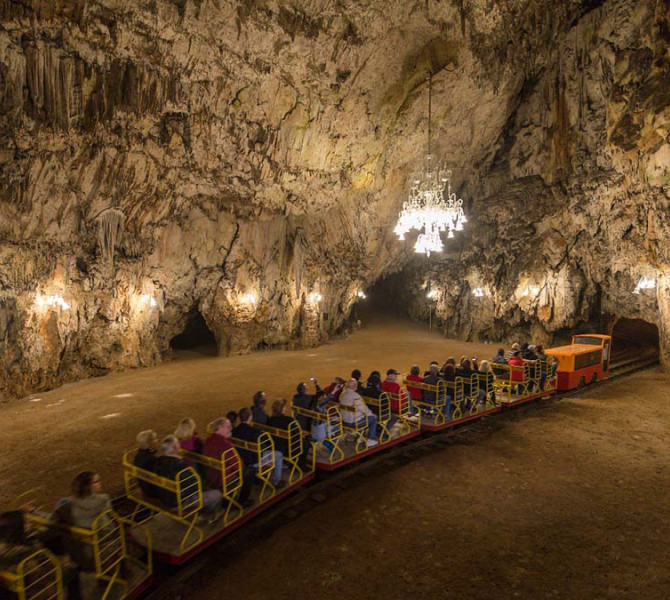 Turisticni vlakec poln obiskovalcev vozi skozi veliko podzemno dvorano Postojnske jame, osvetljeno z lestenci in toplimi lucmi.