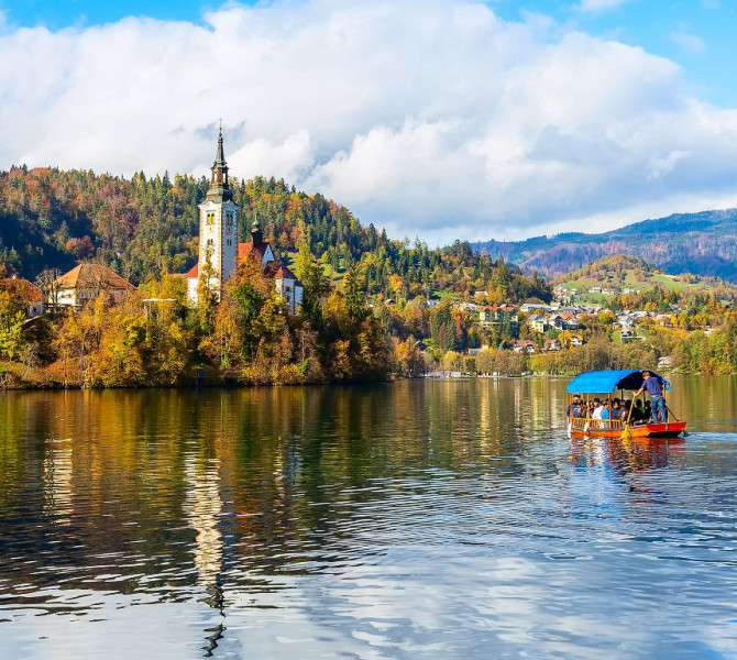 Une barque en bois sur le lac navigue vers l'îlot abritant la petite église. Couleurs d'automne.