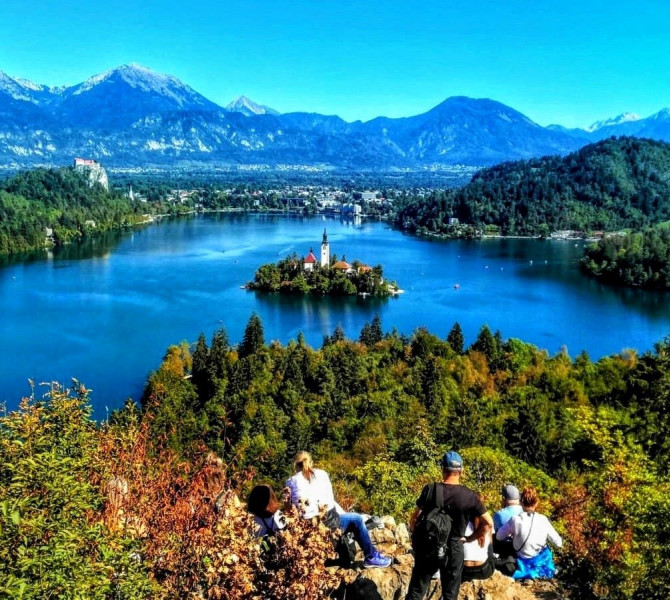 View from a hill of Lake Bled and the island. Four hikers on the hill. Mountains in the background.
