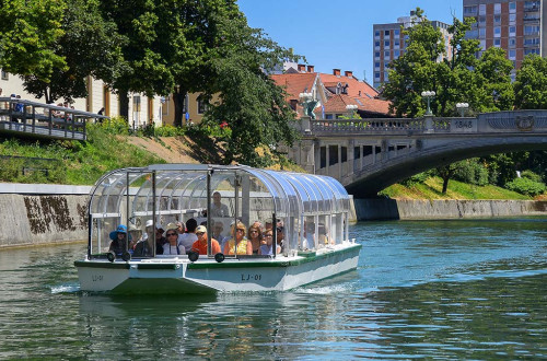 Turisticna ladjica na Ljubljanici. V ozadju narava, most in stolpnica.