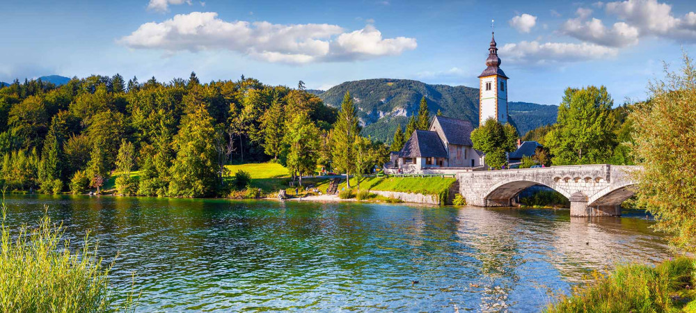 A lake surrounded by a forest in autumn colors. In the background, a bridge and a small church.