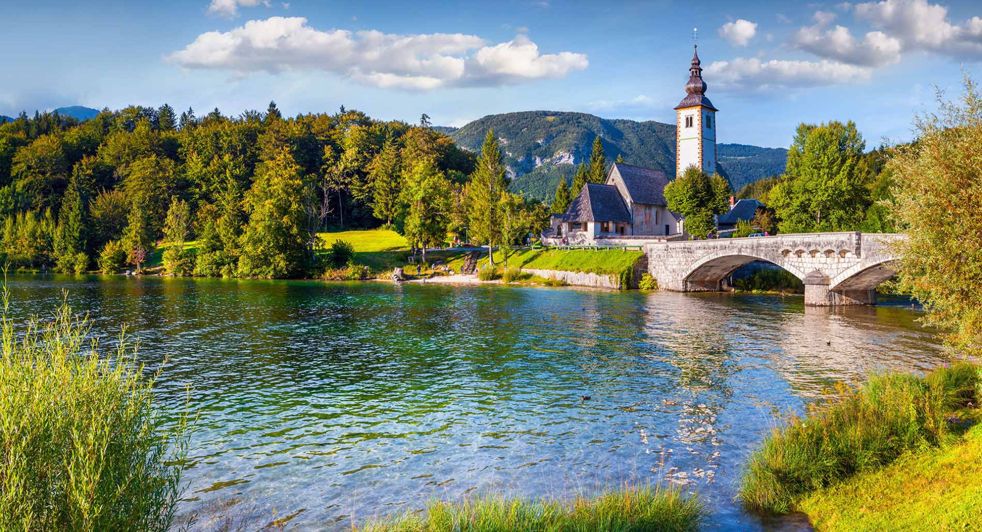 Jezero obdano z gozdom v jesenskih barvah, v ozadju most in cerkvica.