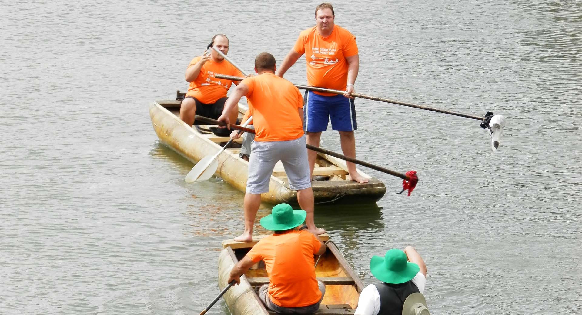 Ljudje so v colnih na reki – nekateri veslajo, drugi pa stojijo in se z vesli med seboj borijo.