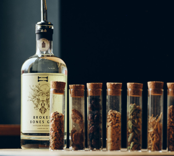 A bottle of gin stands beside a row of small glass test tubes filled with various spices and botanical additions.