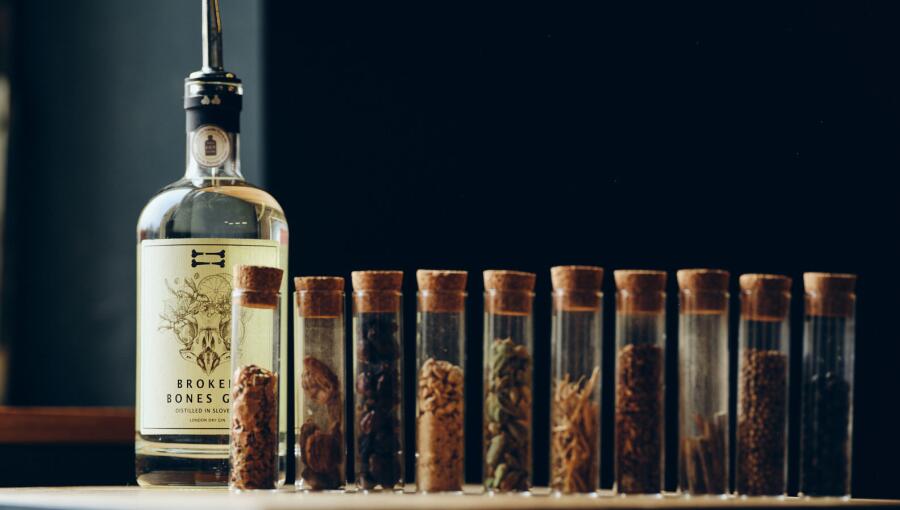 A bottle of gin stands beside a row of small glass test tubes filled with various spices and botanical additions.