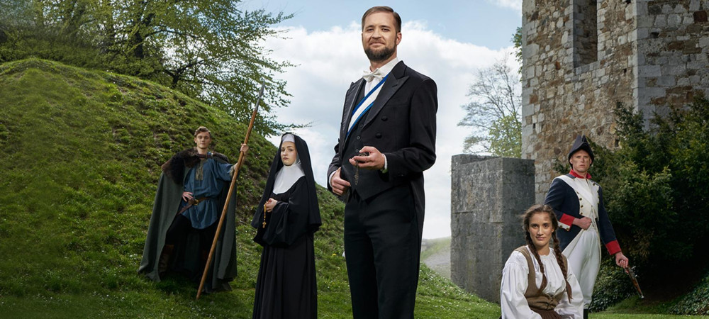 Fünf Personen in Kostümen stehen vor einem Burghügel und einer Mauer.