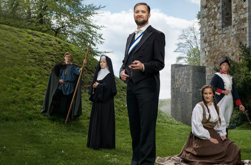 Fünf Personen in Kostümen stehen vor einem Burghügel und einer Mauer.