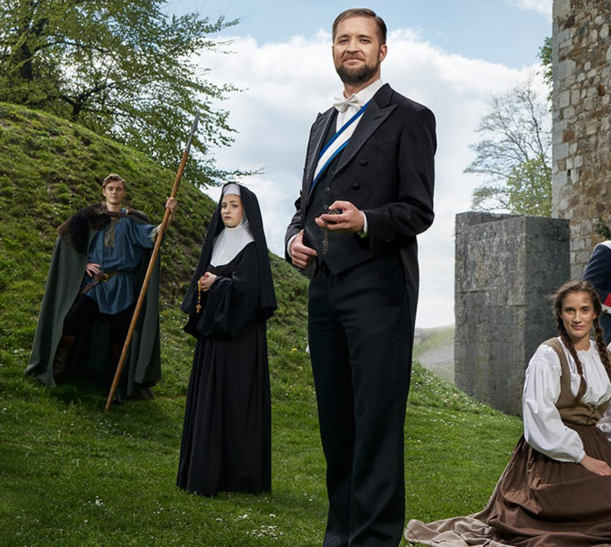 Five people dressed in costumes, standing in front of a castle hill and wall.