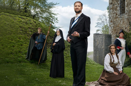 Pet ljudi oblecenih v kostume stoji pred grajskim hribom in zidom.