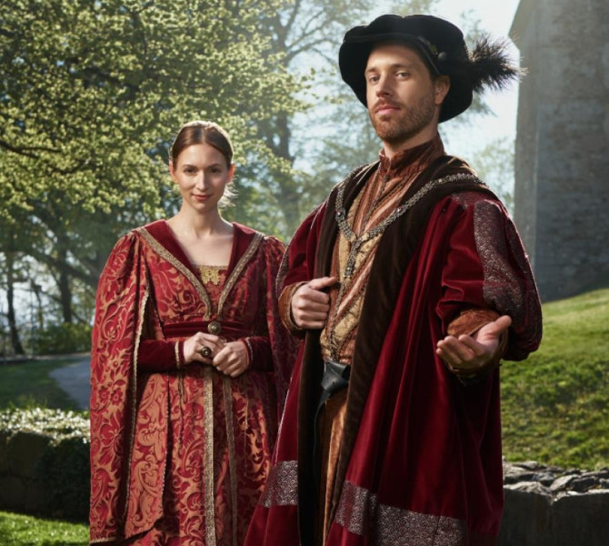 Two people dressed in medieval red costumes, standing beside a castle with trees behind them.