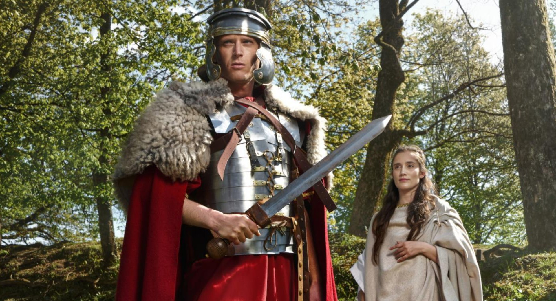 Un homme vêtu d’un costume de chevalier médiéval et une femme en robe médiévale se tiennent parmi les arbres.