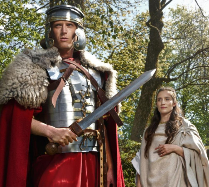 Man dressed in a medieval knight costume and a woman in a medieval dress are standing among trees.