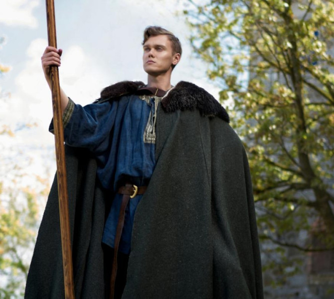 Man dressed in a costume holding a stick, standing among trees.