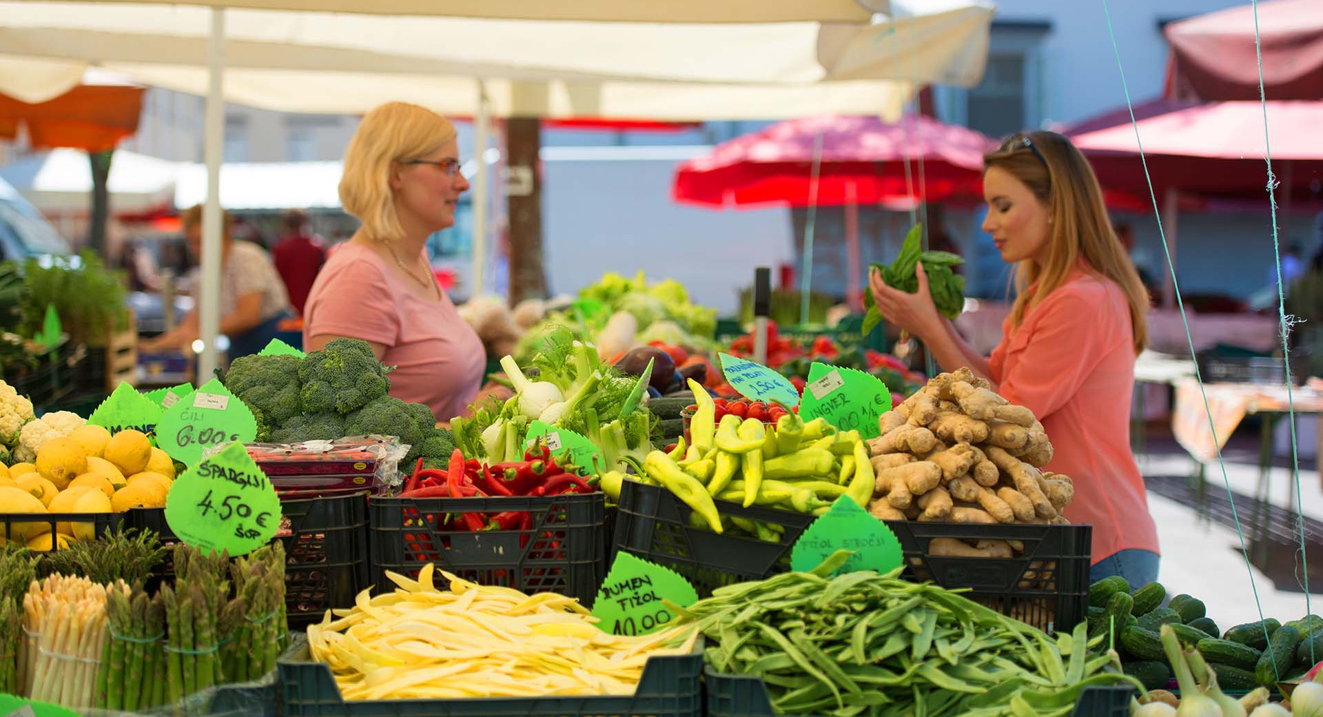 Stojnice na glavni tržnici z različno ponudbo. V ozadju prodajalka in obiskovalka.