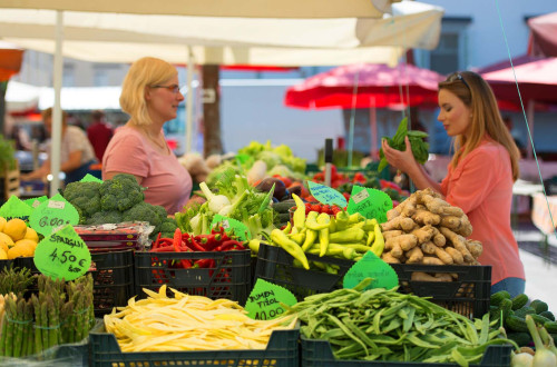 Stojnice na glavni tržnici z različno ponudbo. V ozadju prodajalka in obiskovalka.