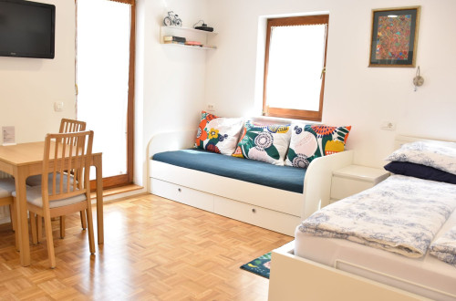 Ein Raum mit einem kleinen quadratischen Holztisch auf der linken Seite, einem weißen Sofa mit einer grün-blauen Decke in der Mitte und einem weißen Bett mit weißer Bettwäsche auf der rechten Seite.
