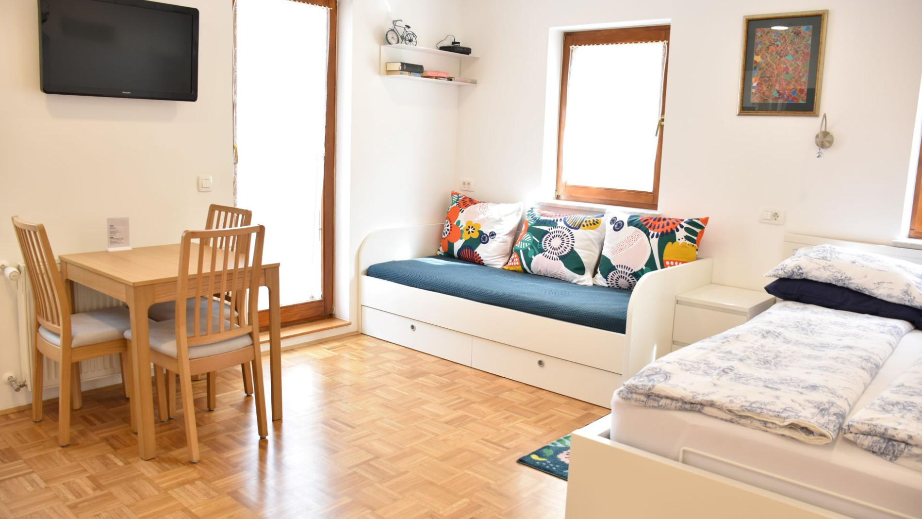 A room with a small square wooden table on the left, a white sofa with a green and blue throw in the centre, and a white bed with white bed linen on the right.