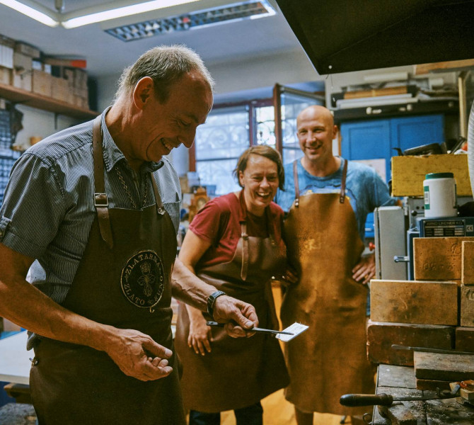 Zwei Workshop-Teilnehmende beobachten den Goldschmied beim Schmieden einer Metallplatte.