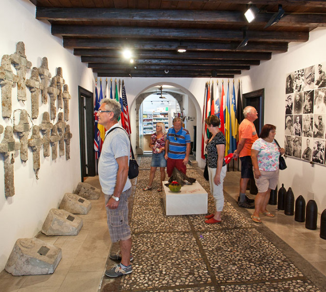 I visitatori del museo osservano una mostra sul Fronte Isontino, che include croci di pietra sulla parete, ritratti di soldati in bianco e nero e proiettili d'artiglieria sul pavimento.