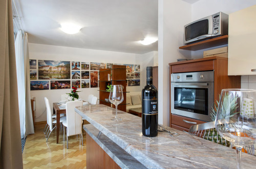 Kuhinja s šankom, na katerem je steklenica vina in kozarca,. V ozadju je jedilna miza s stoli in na njeni desni kavč. 
