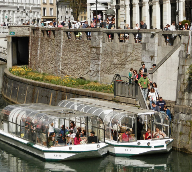 Two tourist boats with passengers on the Ljubljanica River. In the background, the riverbank and the Triple Bridge.