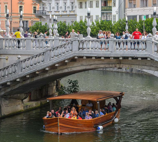 A wooden boat, full of people, is sailing on the river, with a stone bridge above and people on it.