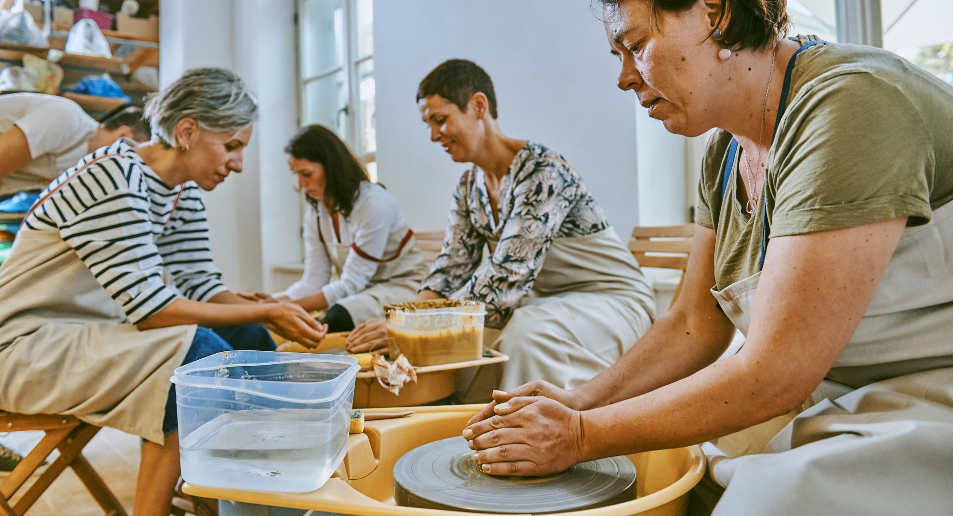 Udelezenke oblikujejo glino na delavnici za loncarstvo, kjer skupaj ustvarjajo na vrtecih kolesih.