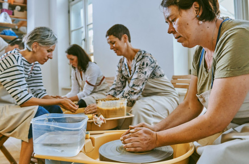 Udelezenke oblikujejo glino na delavnici za loncarstvo, kjer skupaj ustvarjajo na vrtecih kolesih.