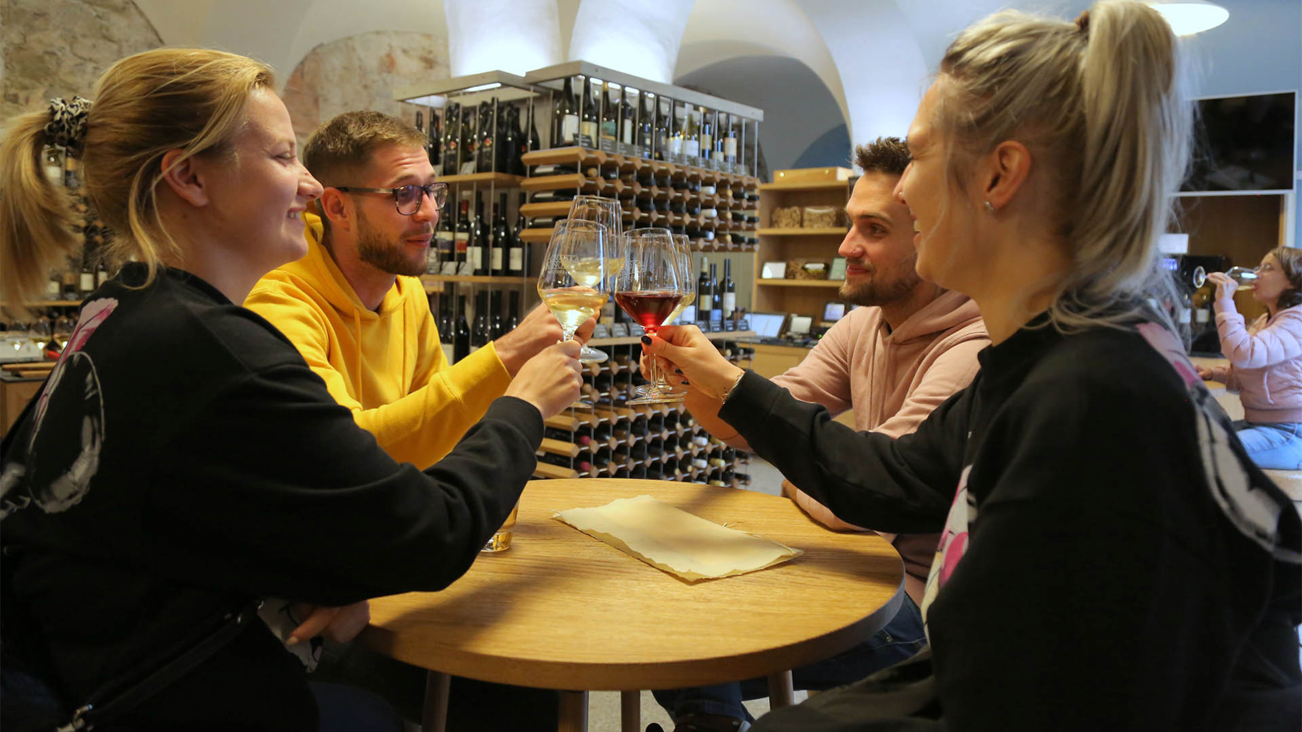 Štirje prijatelji nazdravljajo s kozarcem vina. V ozadju vinska vitrina. 
