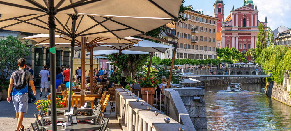 Tables, umbrellas, and chairs on the riverbank. In the background, a bridge and a church.